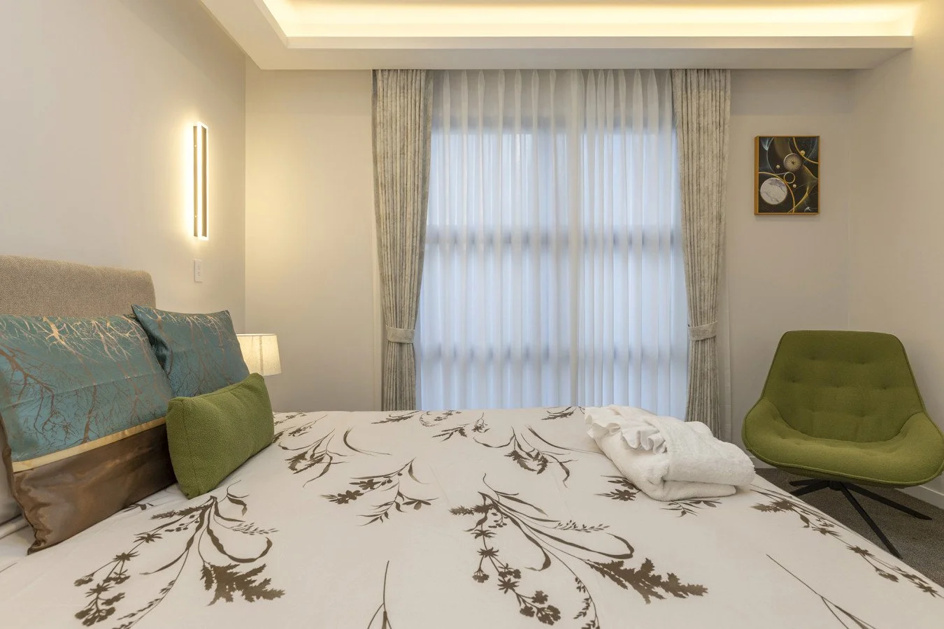 A bedroom with a bed covered in white and brown floral bedding, a beige headboard, blue and green pillows, a small lamp on bedside table, beige curtains, a green armchair, and modern wall art.