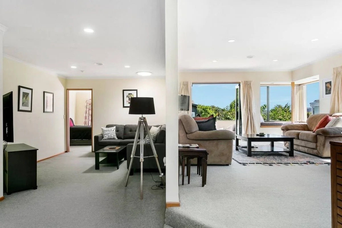 A split view of two living rooms, with the left room having dark furniture, a TV, and framed pictures, and the right room featuring light-colored sofas, large windows with curtains, and a view of trees outside.
