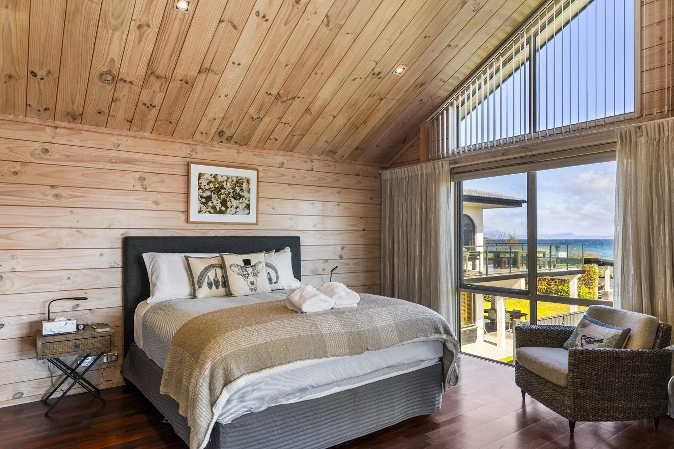Bedroom with wood-paneled vaulted ceiling, large window and sliding glass door showing ocean view, bed with pillow decorated with a deer wearing a hat, armchair with a pillow, and outdoor balcony.