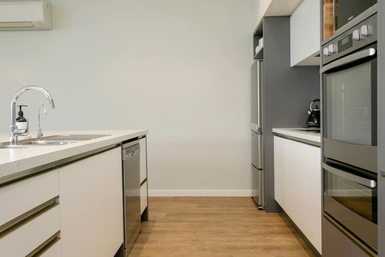 Minimalist modern kitchen with white cabinets, stainless steel appliances, wooden floor, and a white countertop with a sink and soap dispenser.
