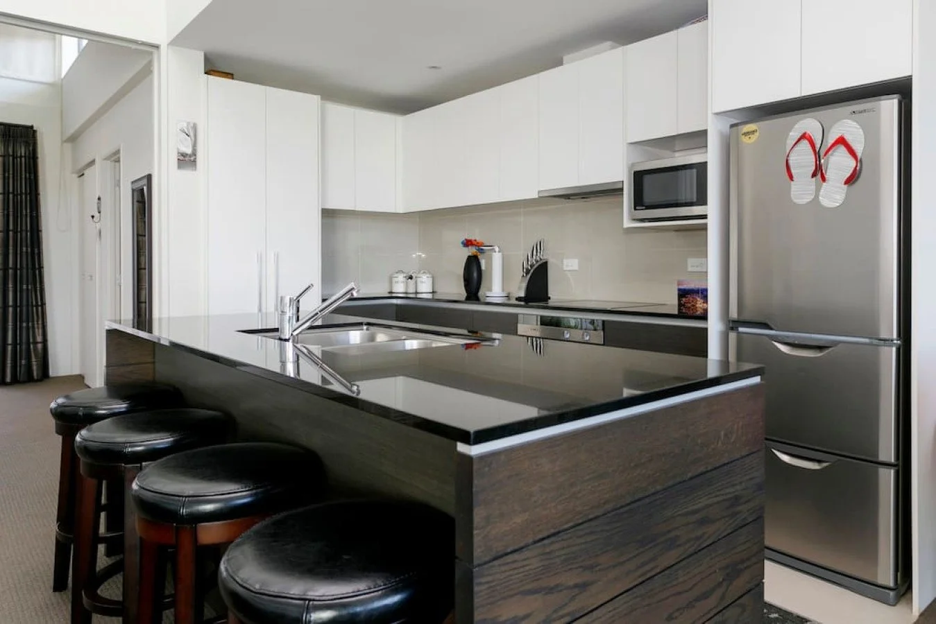 Modern kitchen with black countertop island, four black bar stools, white cabinets, stainless steel refrigerator with flip-flops magnet, microwave, and various small kitchen appliances.
