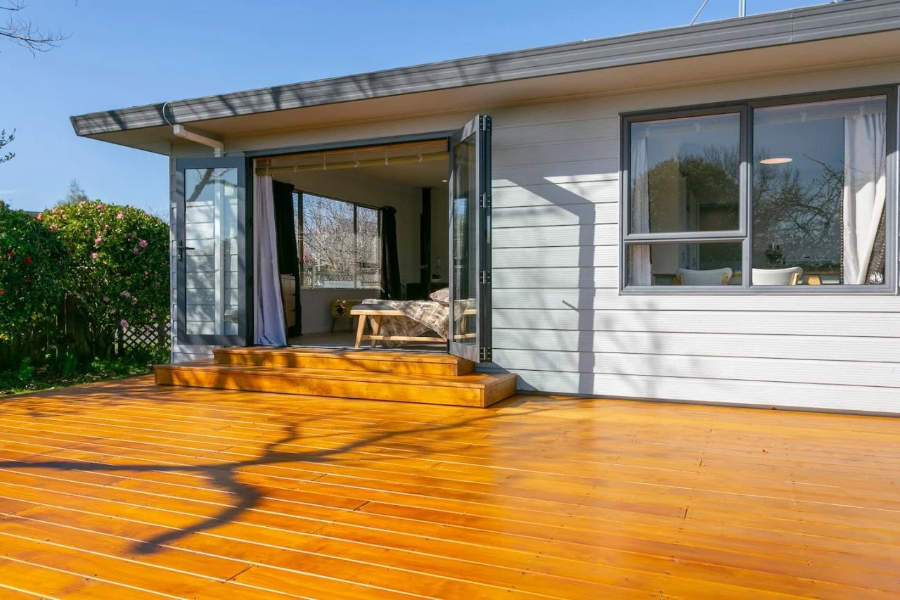 A view of a house exterior with a wooden deck and open glass door leading to a bedroom with a bed and window.