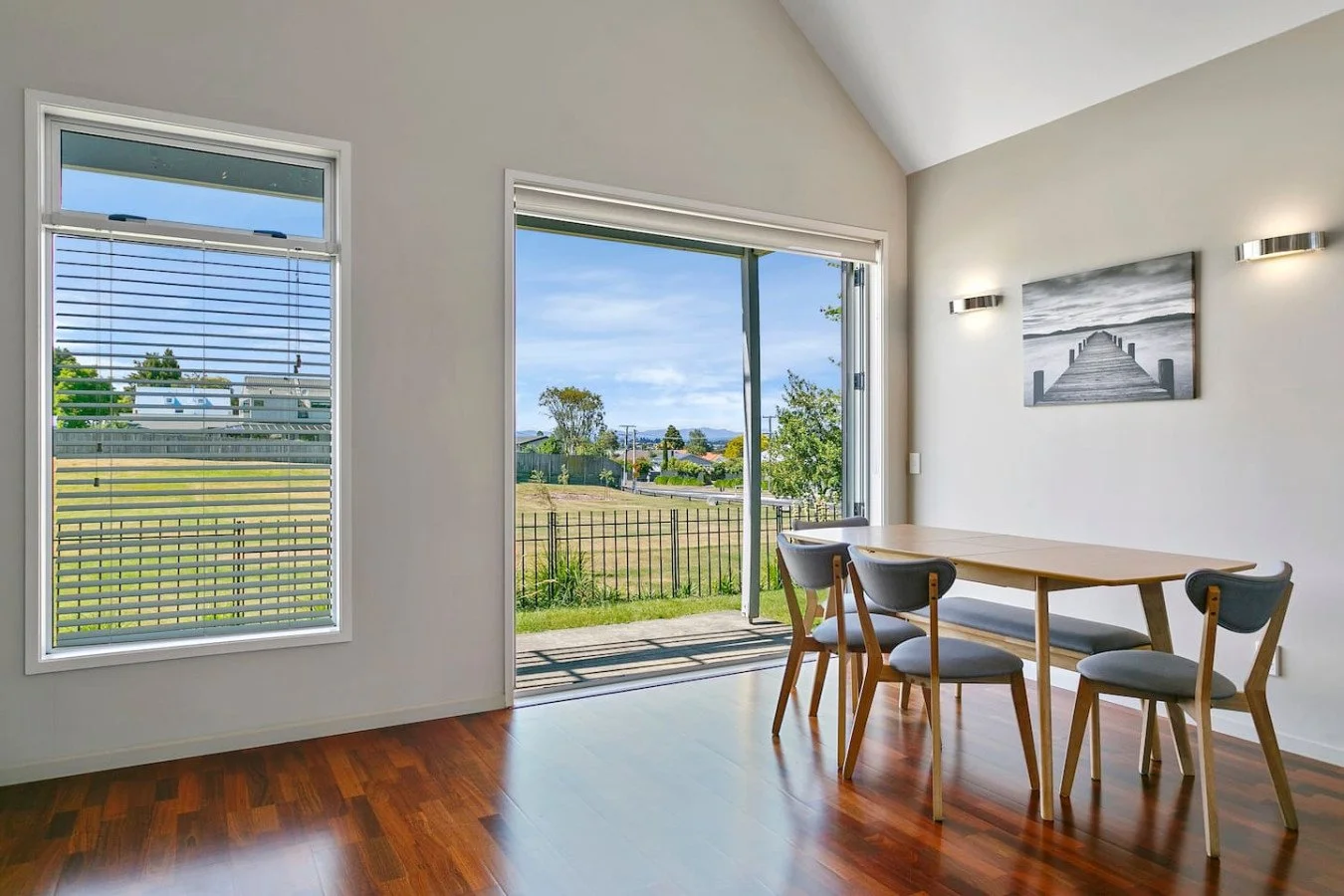 Dining area with wooden table and four chairs, sliding glass door open to backyard, and window with blinds showing outdoor scenery.
