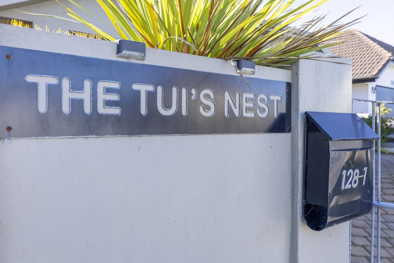 Street sign reading 'The Tui's Nest' on a concrete wall with a black mailbox labeled '128-1' and a plant with long, pointed leaves behind the sign, in a residential neighborhood.
