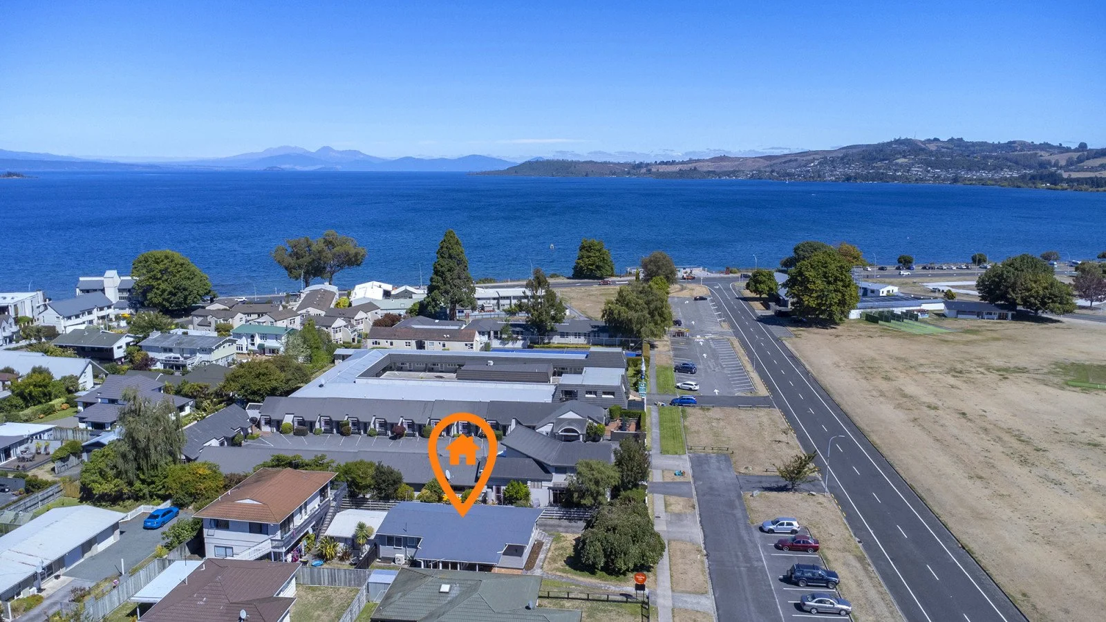 Aerial view of a lakeside neighborhood featuring various residential houses, a parking lot, trees, and an adjacent open field, with a large body of water and distant hills or mountains in the background.