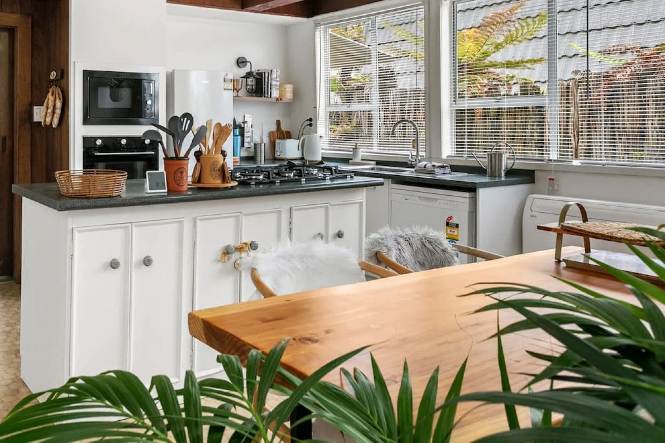 A cozy kitchen with white cabinets, black countertops, and stainless steel appliances. There's a wooden dining table in the foreground with green plants nearby. Large windows with blinds let in natural light, and various kitchen utensils and applianc
