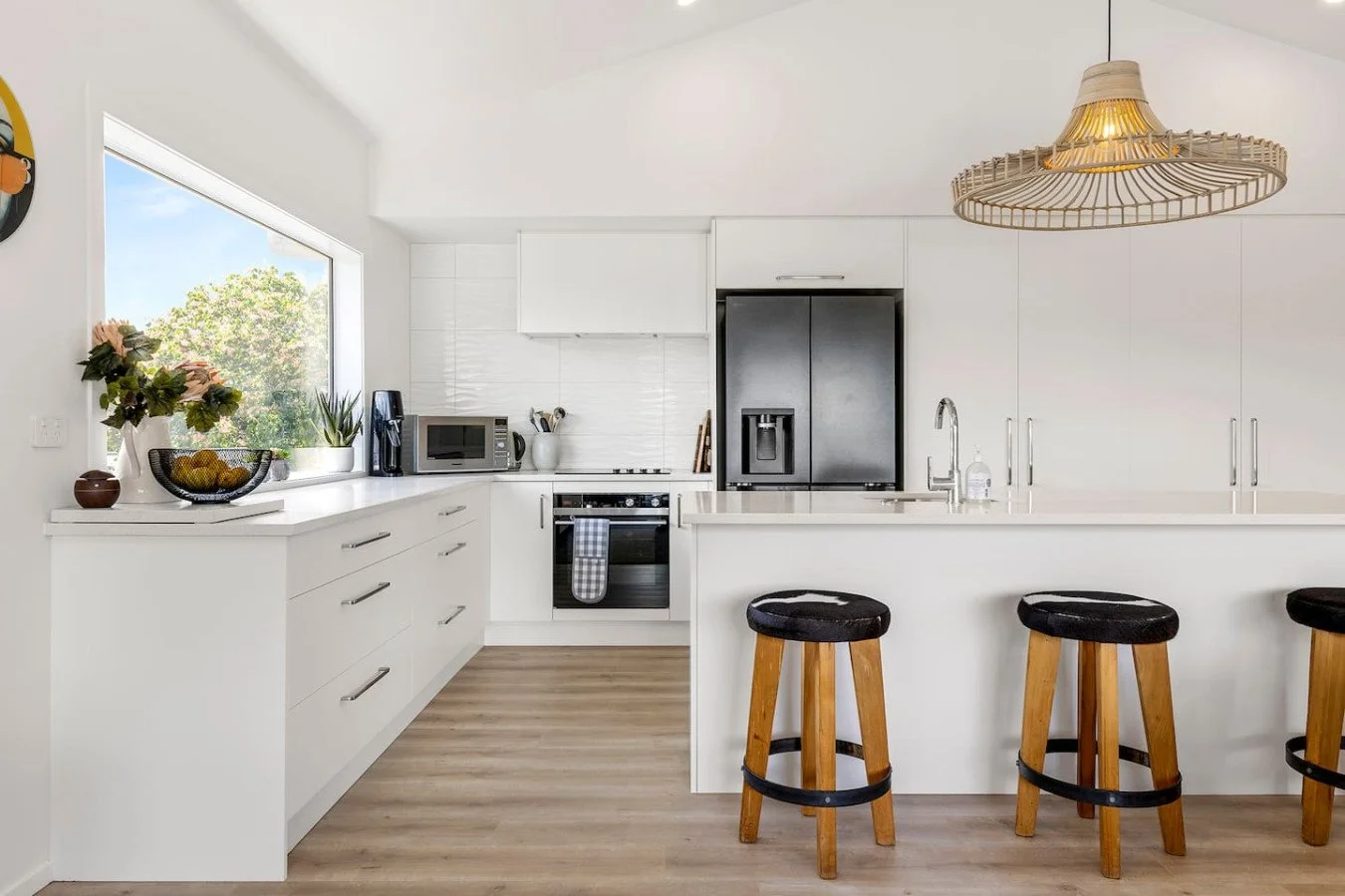 Modern white kitchen with wood bar stools, a black refrigerator, microwave, and window view of trees.