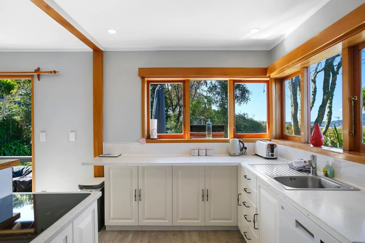 Kitchen with white cabinets, wooden window frames, a sink, and a view of trees outside.