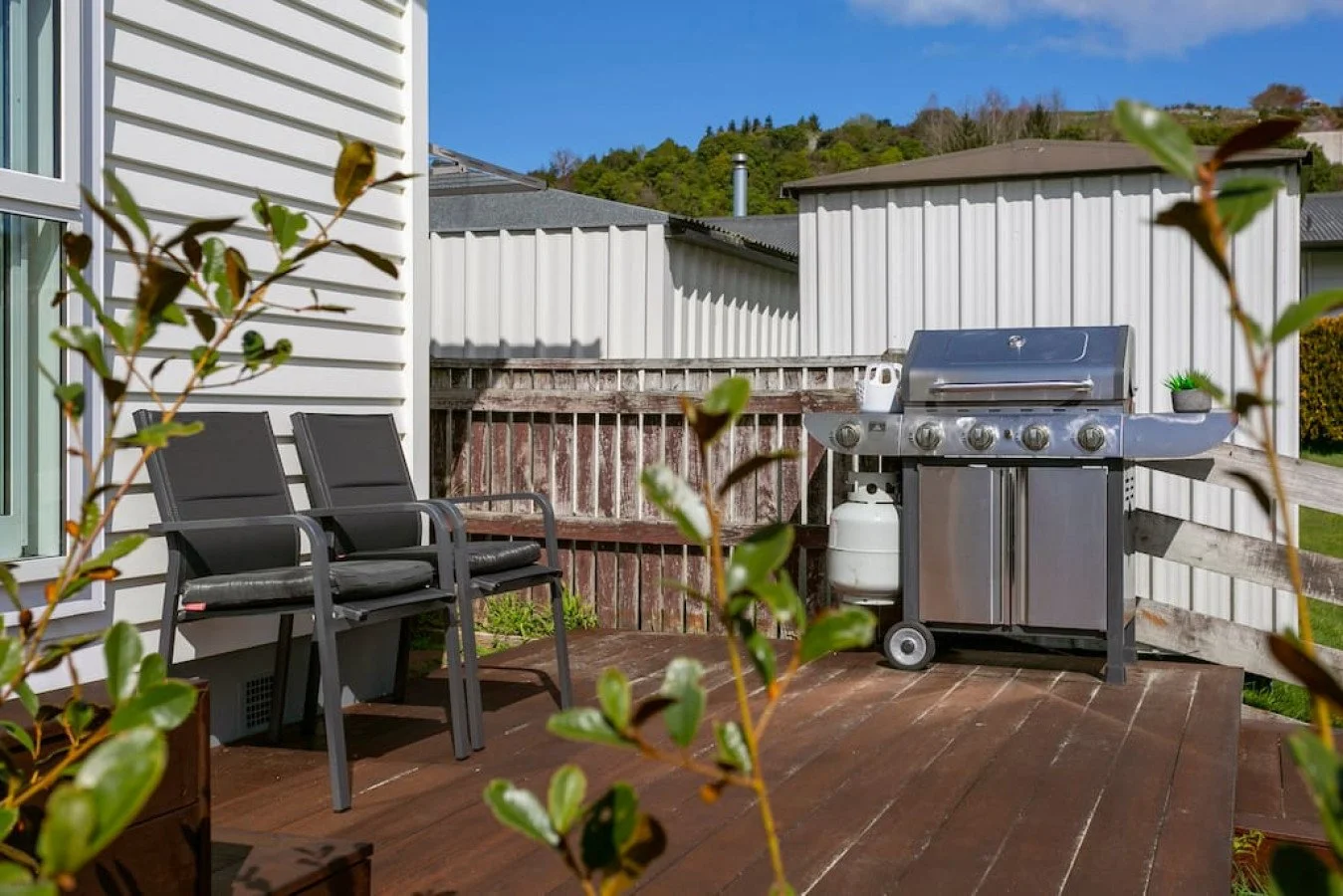 Backyard deck with two black chairs, a stainless steel gas grill with propane tank, and some potted plants, enclosed by wooden and metal fencing, under a blue sky.