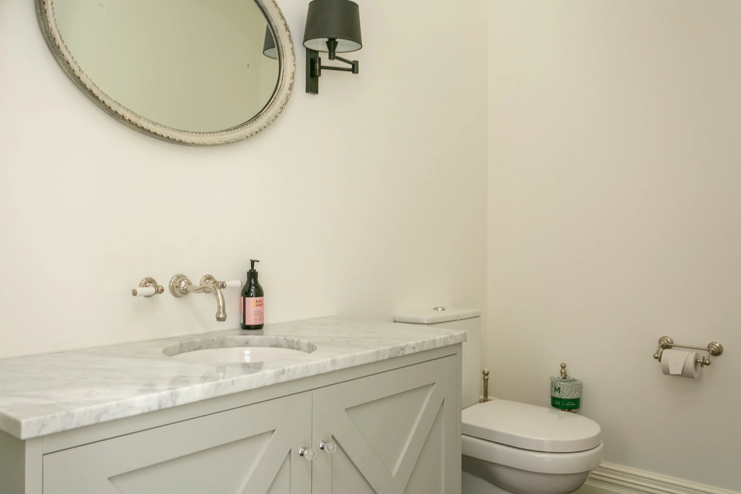 A minimalist bathroom featuring a white vanity with a marble countertop, a mounted oval mirror, wall-mounted lamps, a soap dispenser, a toilet with a roll of toilet paper on the holder, and a green toilet paper roll cover.