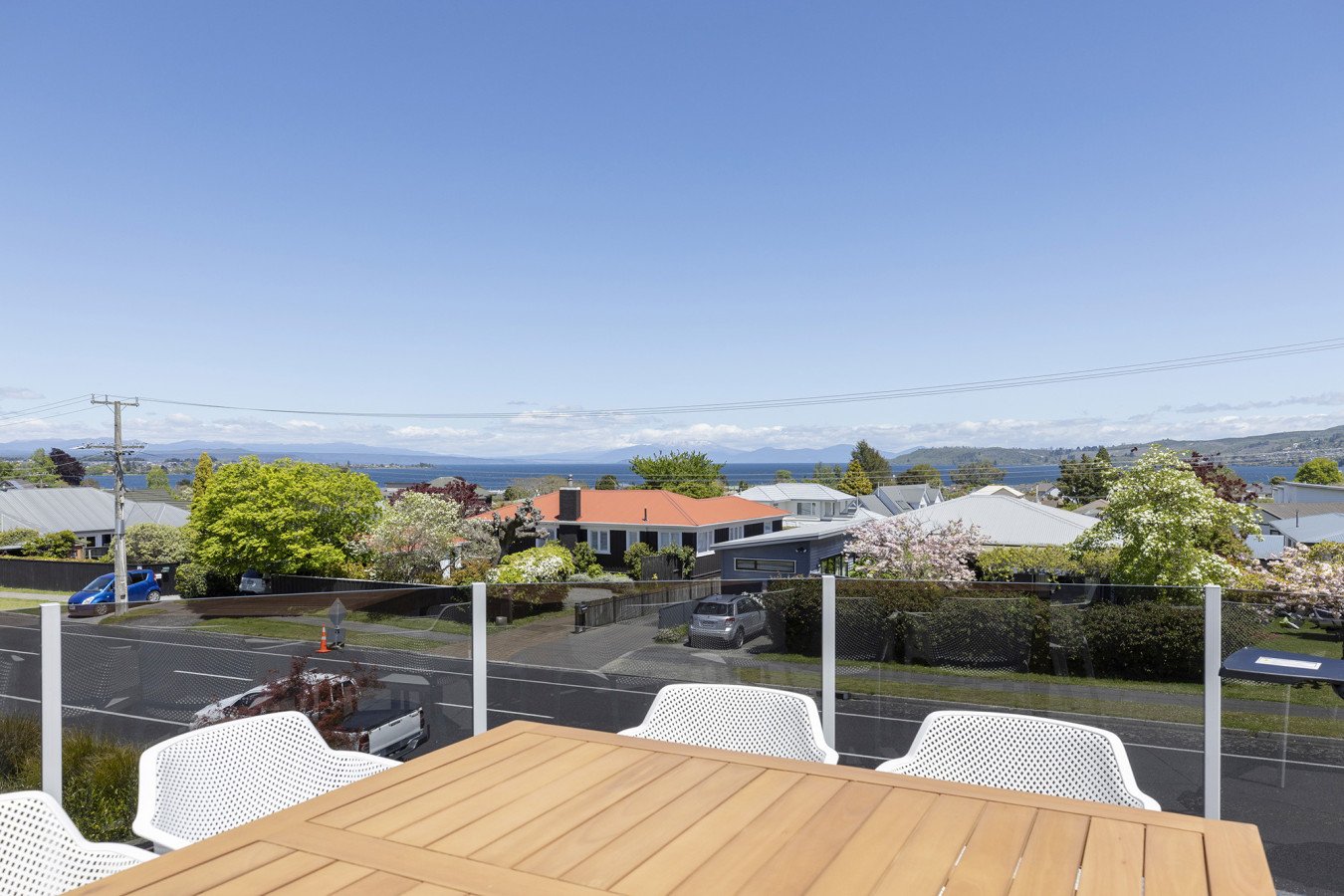A view from a balcony with a wooden table and white chairs overlooking a suburban neighborhood with houses, trees, and a body of water in the background under a clear blue sky.