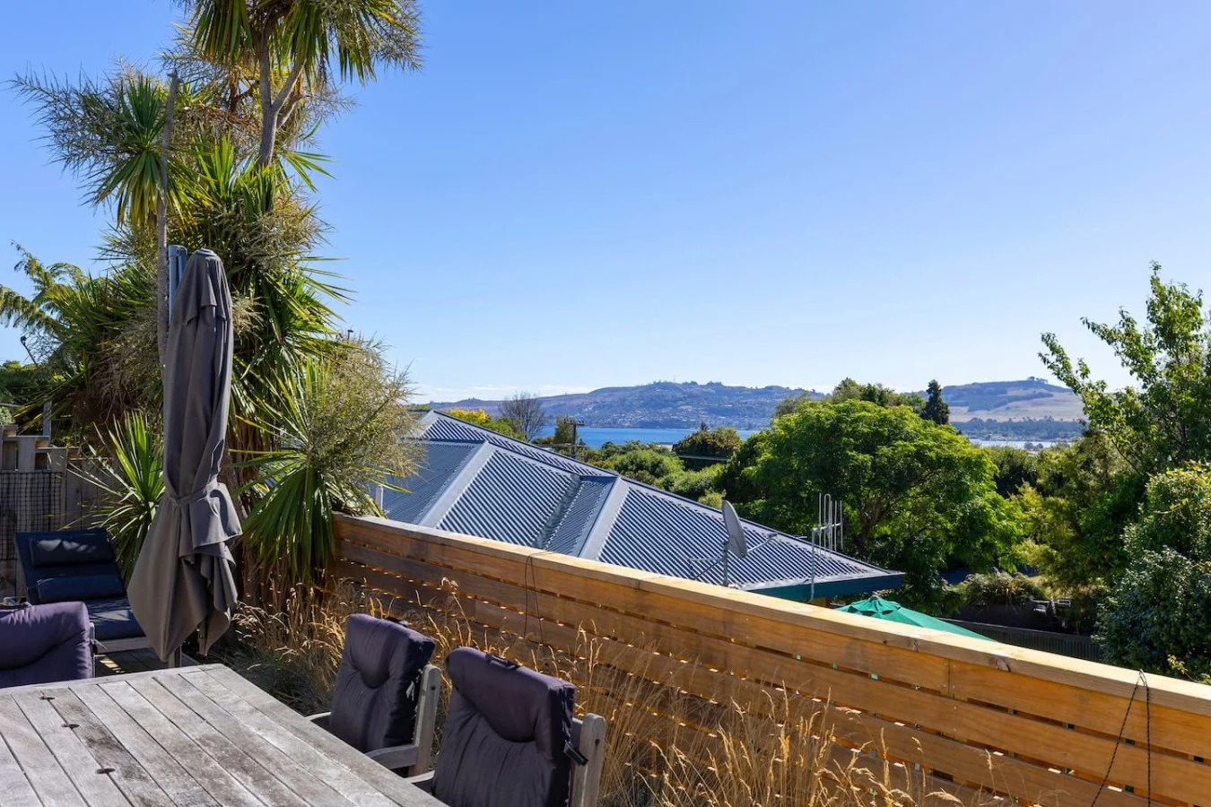 Outdoor deck with wooden railing, chairs, table, a large umbrella, trees, rooftops, and a distant view of a body of water and hills under a clear blue sky.