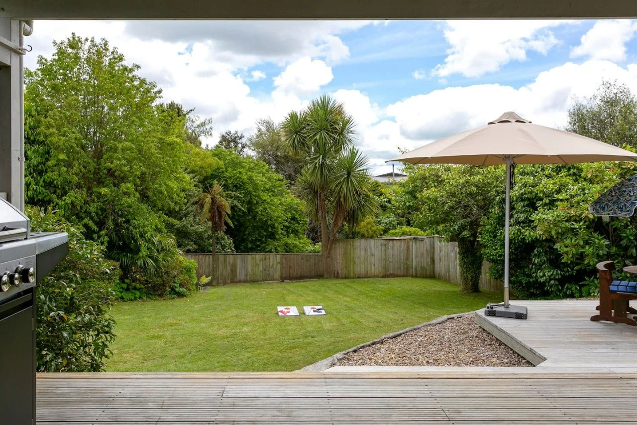 A backyard with a grassy lawn, surrounded by trees and shrubs, a wooden fence, a deck with an umbrella and lounge chair, and two cornhole boards on the grass.
