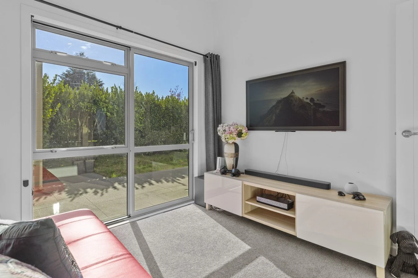 Living room with sliding glass door, view of green trees outside, wall-mounted TV with mountain scene, white and wood TV stand, vase with flowers, soundbar, remote controls, small decorative object, and plush dog toy on beige carpet.