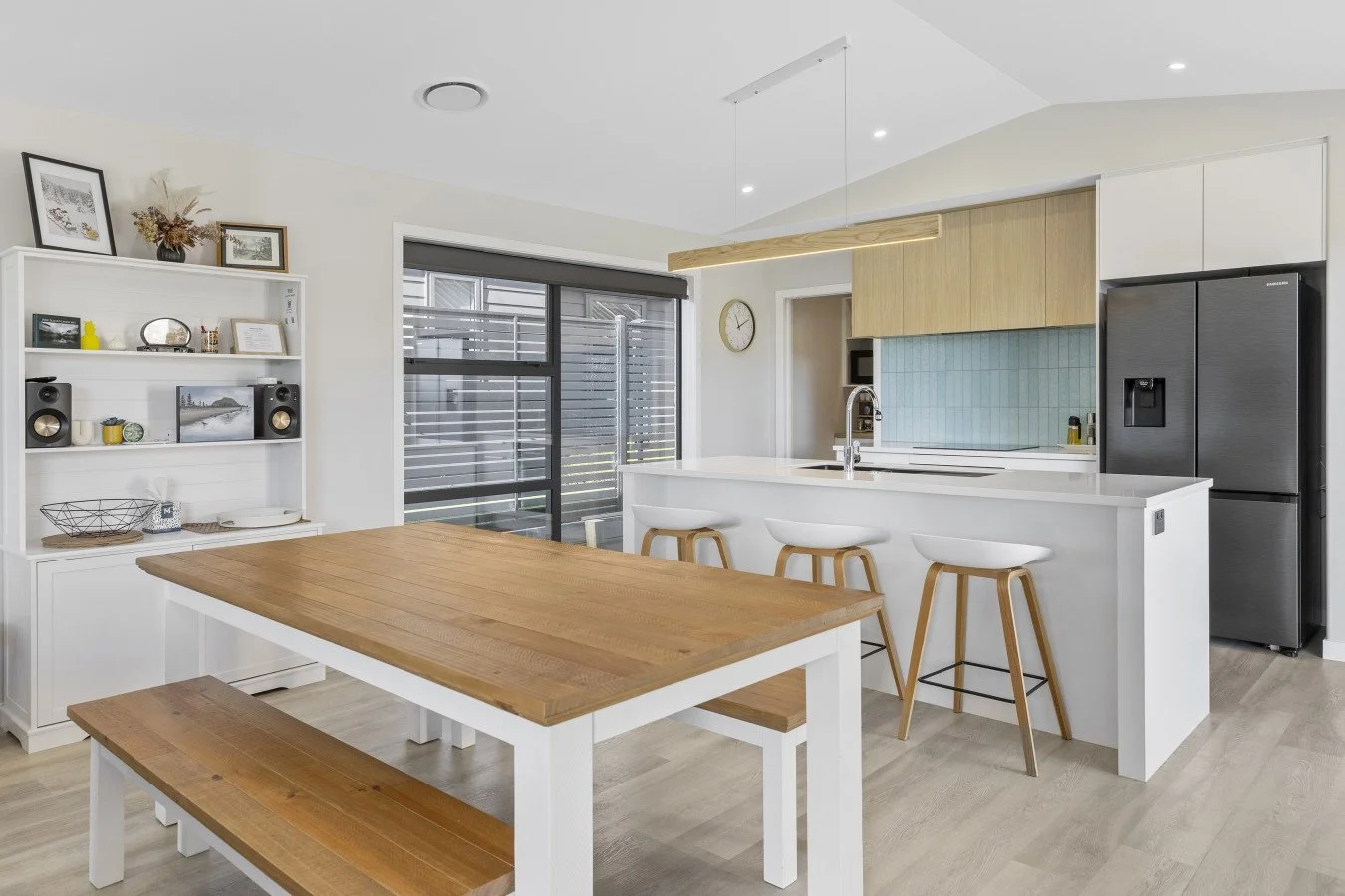 Modern kitchen with white cabinets, a wooden dining table with matching benches, and a breakfast bar with three white stools. Large window with blinds, view of outside patio. Black refrigerator, wall clock, and a white shelving unit with decor and bo
