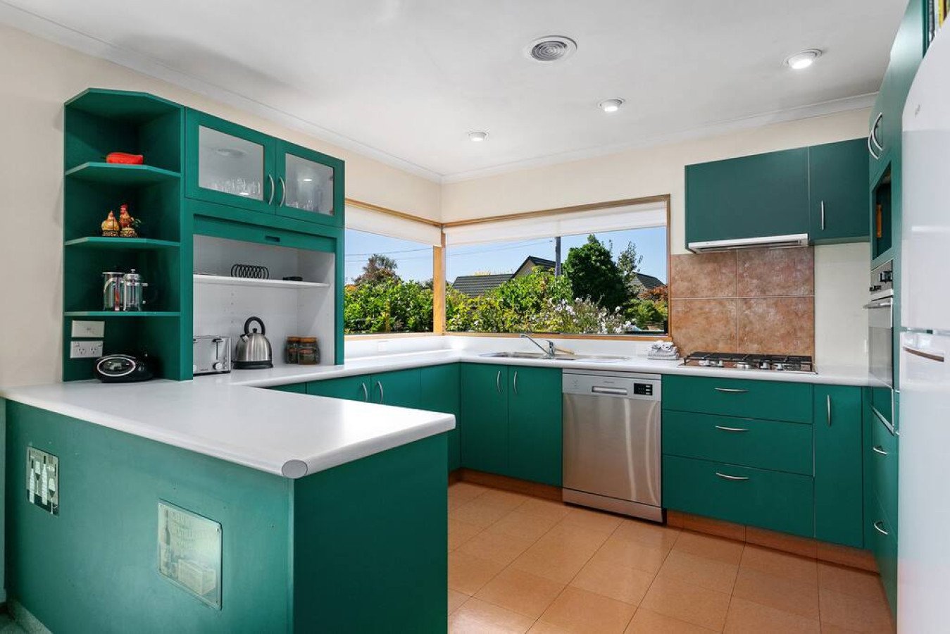 Kitchen with green cabinets, white countertops, a window with a view of trees and houses, stainless steel appliances, including a dishwasher and oven, and a tiled backsplash.
