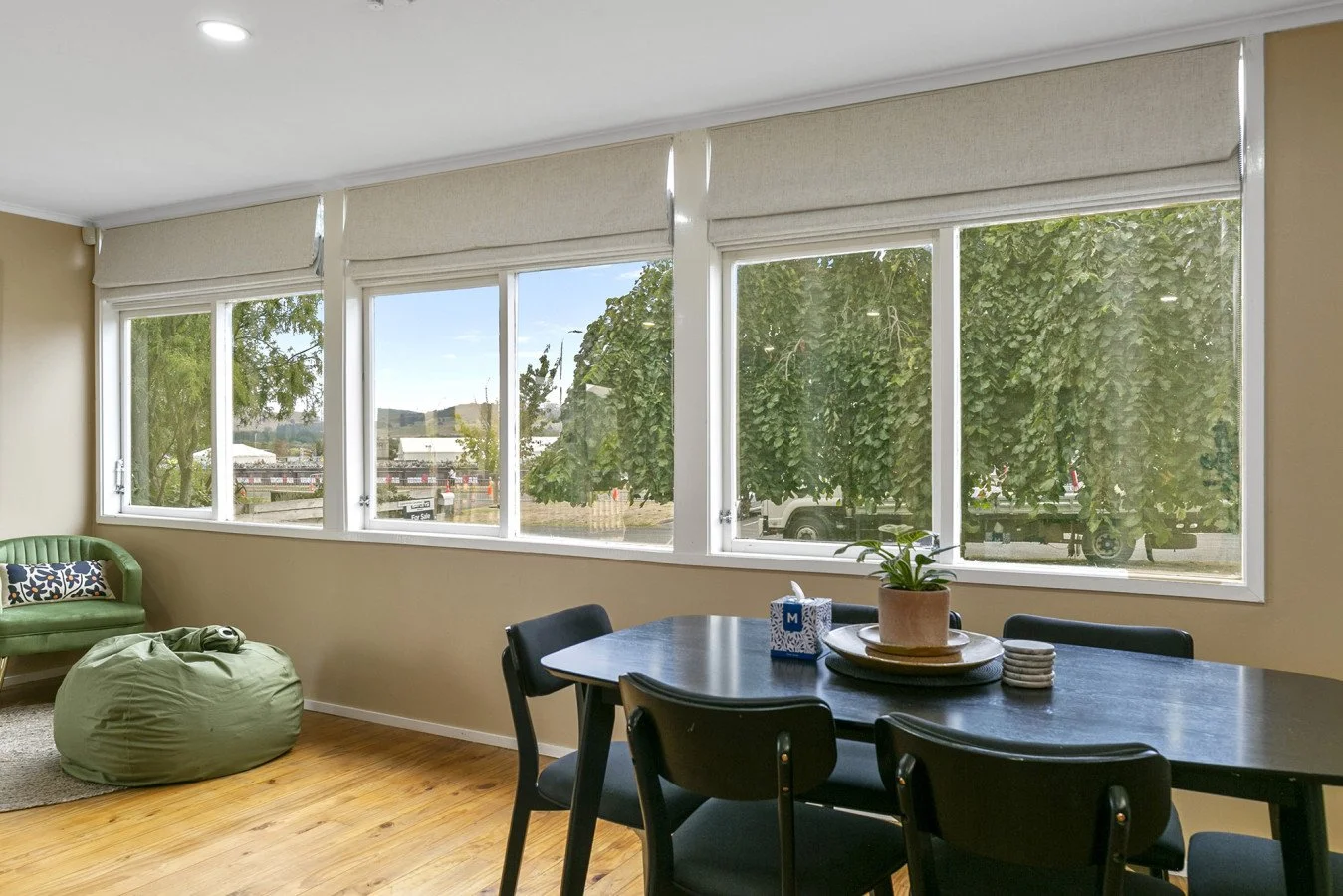 Interior view of a room with large windows, a black dining table with chairs, a potted plant, a green bean bag, and a green vintage-style sofa, with trees and parked vehicles outside.
