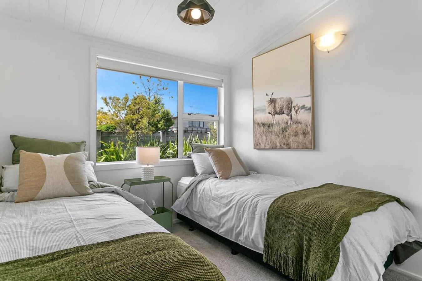 A cozy bedroom with two single beds, white bedding, green blankets, and decorative pillows, a window showing trees outside, and a wall art of a cow in a field.