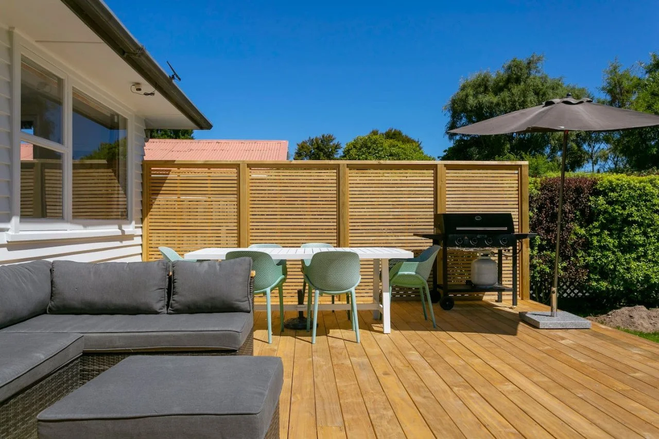 Outdoor patio with gray sectional sofa, white dining table with chairs, barbecue grill, large umbrella, wooden deck, garden hedges, and blue sky.