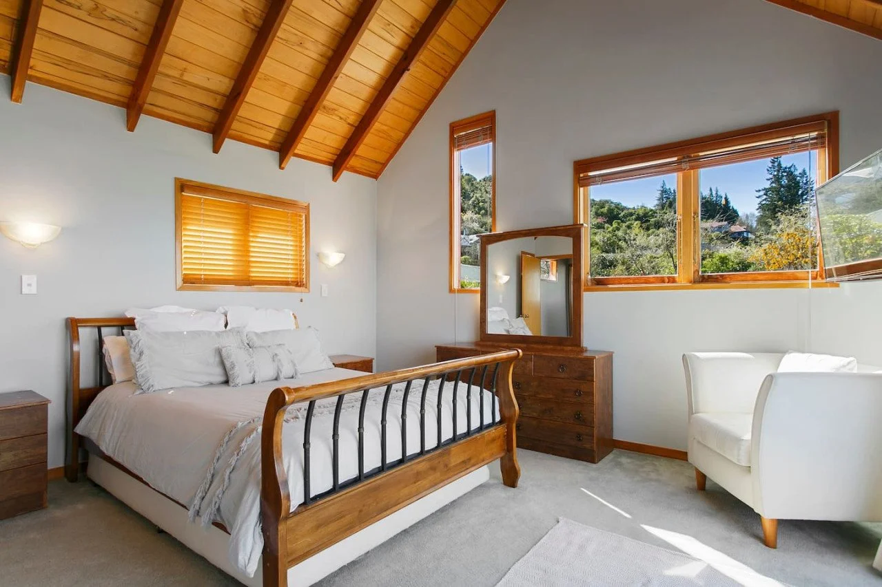 A bedroom with a wooden vaulted ceiling, a bed with white bedding and pillows, a white armchair, a wooden dresser with a mirror, and windows showing greenery outside.