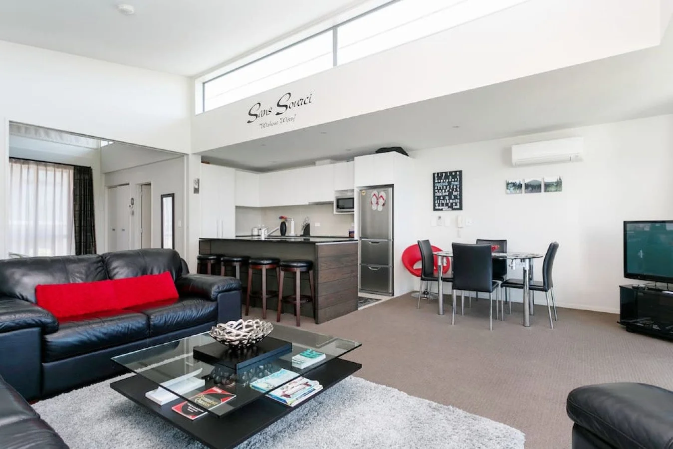 Living room with black leather sofas, glass coffee table, and a TV. An open kitchen with a breakfast bar, and a dining area with four chairs. Bright space with white walls, large windows, and modern decor.