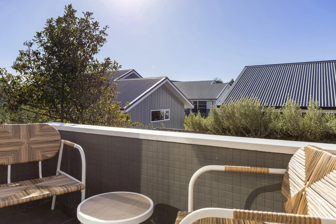 A balcony with two wicker chairs and a small wicker table, overlooking neighboring houses and trees under a clear blue sky.