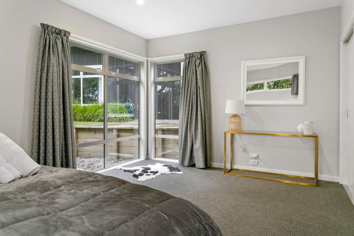 Modern bedroom with a sliding glass door, gray curtains, a cowhide rug, a gold console table with a lamp and decorative items, mirror, bed with gray bedding, and a view of outdoors.
