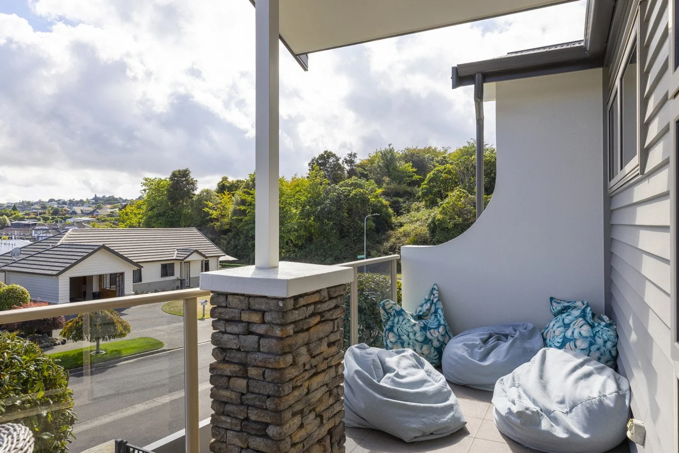 A balcony overlooking a suburban neighborhood with houses, trees, and a hill in the background. The balcony has three large white bean bag chairs and blue patterned pillows, with a glass railing and a stone pillar.
