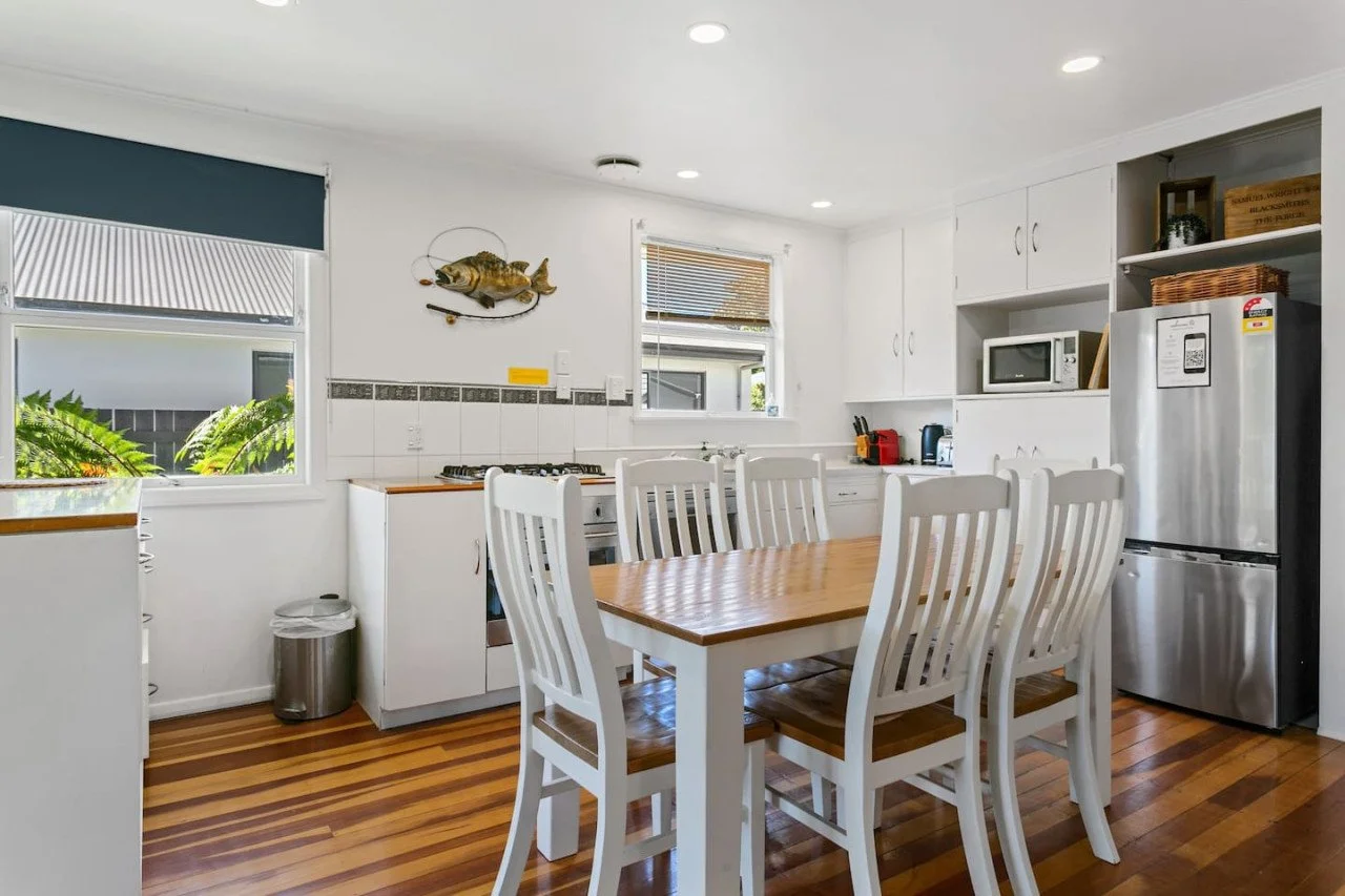 Kitchen with white cabinets, wooden dining table with six chairs, stainless steel refrigerator, microwave, and small appliances on countertop. There are windows with blinds and a fish wall decoration.