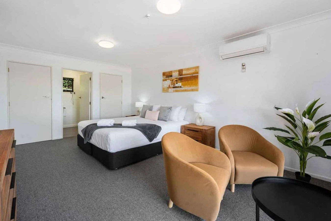 A modern bedroom with a large bed, two tan armchairs, a wooden side table with a lamp, a large plant, and a wall-mounted air conditioner.