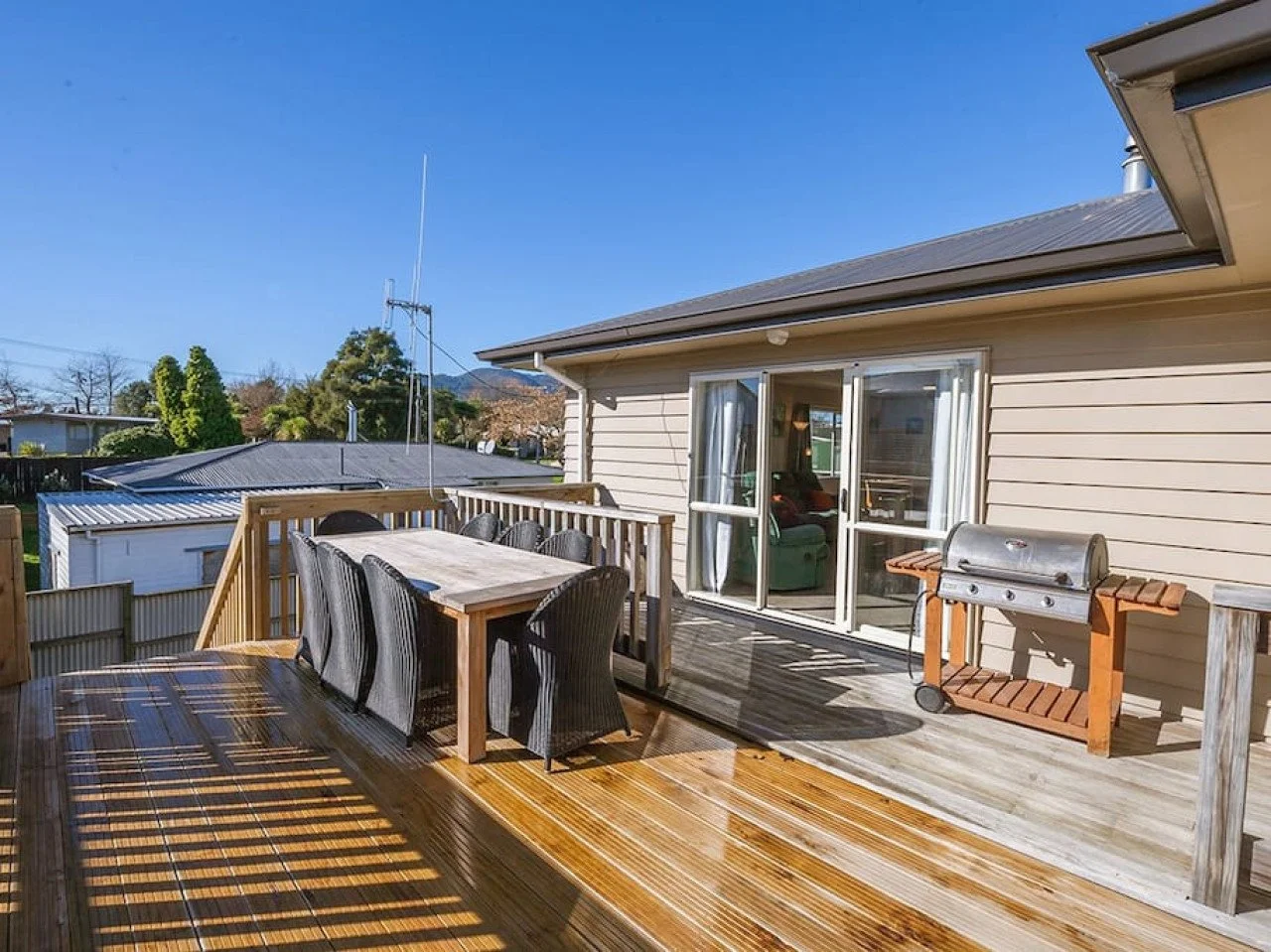 Backyard deck with a dining table and chairs, a barbecue grill, and sliding glass doors leading into the house, under a clear blue sky.