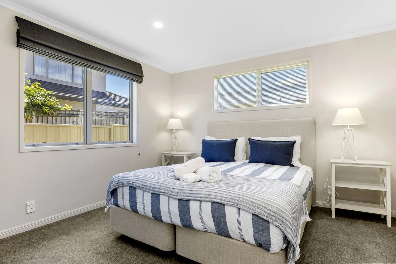 A neatly made bed with white and blue striped bedding, two rolled white towels, and two dark blue pillows in a bright bedroom with two windows, white nightstands, and lamps.
