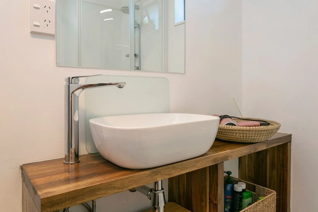Bathroom vanity with a modern bowl sink, chrome faucet, mirror, and a woven basket holding hair styling tools, with a wooden cabinet below containing toiletries.