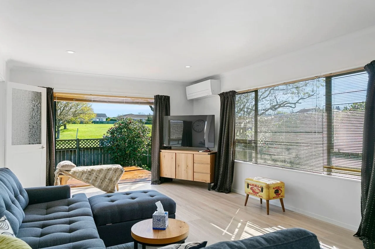 Living room with large windows, a gray sectional sofa, a round coffee table with tissues, a wooden TV stand with a flat-screen TV, and an open door leading to a porch with a view of a grassy yard and trees.