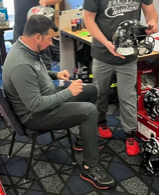 A man sitting on a black folding chair, using a smartphone at an electronics or tech event, with another person standing nearby holding a bicycle or motorcycle helmet.
