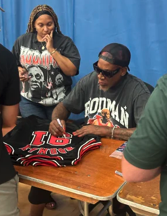 A woman in a graphic T-shirt stands next to a man in sunglasses and a backward cap who is signing or writing on a sports jersey at a table. A person with a green shirt is partially visible nearby.
