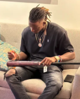 Man with dreadlocks wearing a black t-shirt and ripped jeans, sitting on a beige chair, reading a magazine.