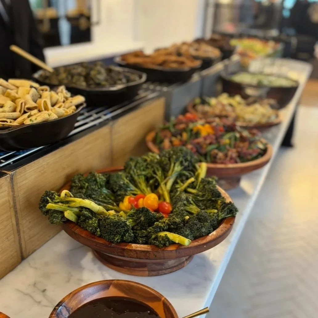 A variety of dishes displayed on a buffet table, including a bowl of cooked broccoli and cherry tomatoes, along with other bowls containing different types of food.