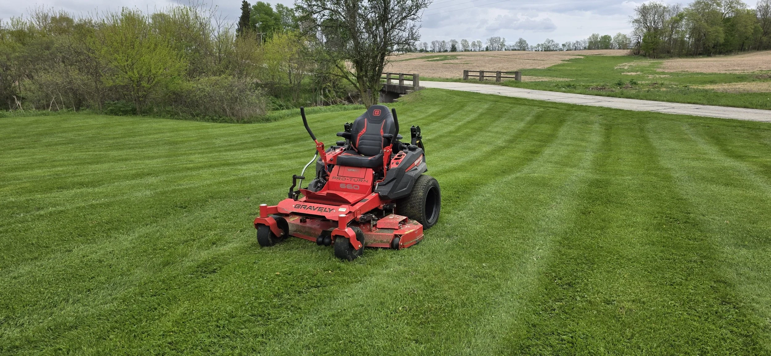 A red zero-turn lawn mower on a green, freshly mowed lawn with striped grass pattern, near a bridge and trees in the background.