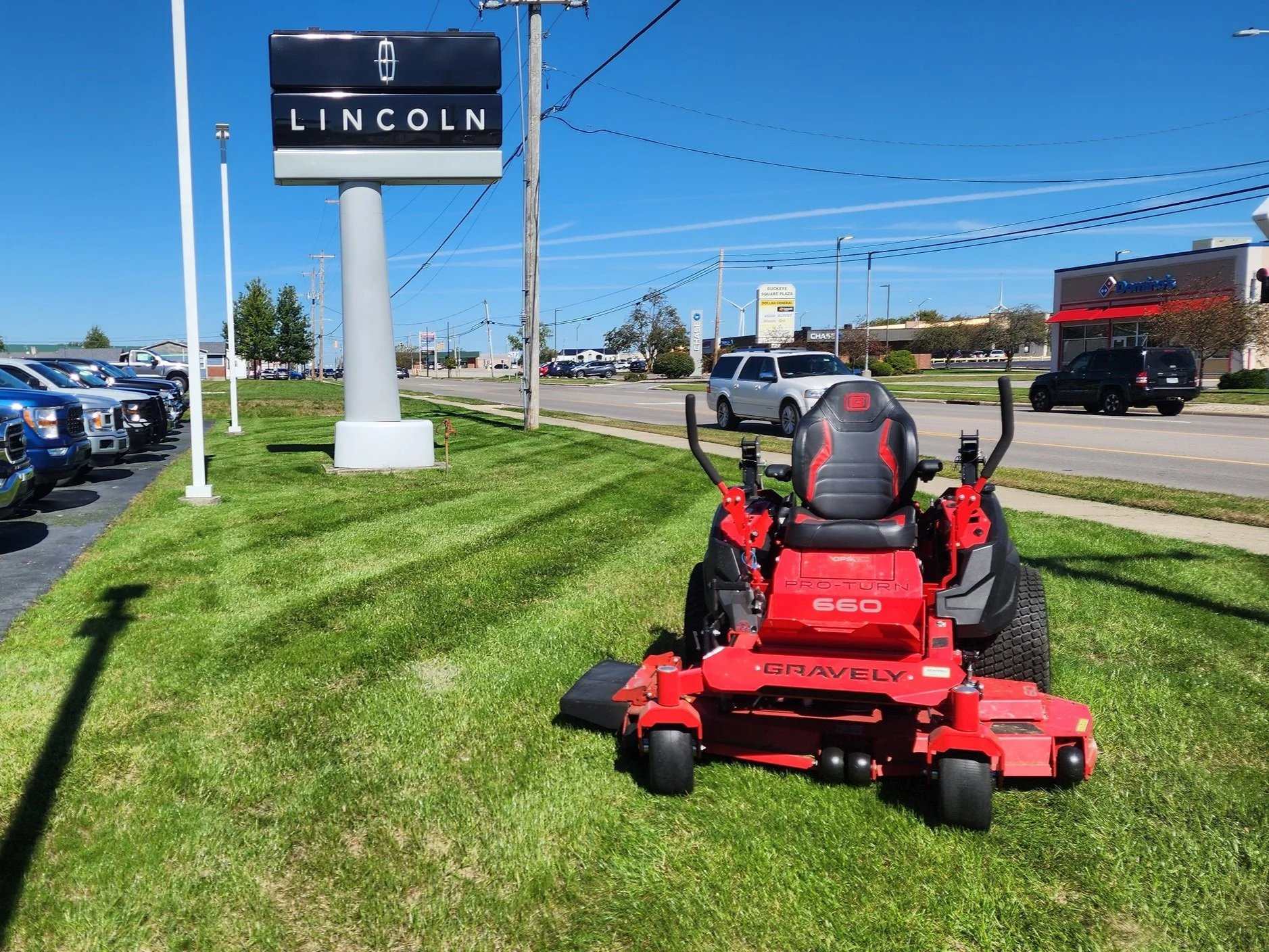 Red commercial mower on freshly cut grass in front of Lincoln dealership with parking lot and cars, and busy street with cars and storefronts in the background.