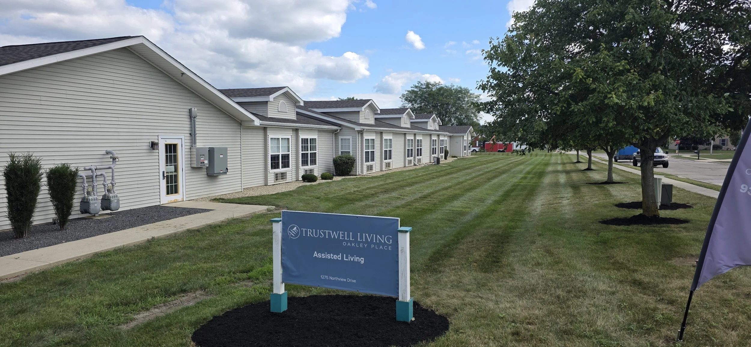 Exterior view of a senior living facility with a grassy lawn, trees, and a sign that reads "Trustwell Living Oakley Place Assisted Living, 1275 Northview Drive." The building has white siding, multiple windows, and small bushes. There are utility boxes and pipes on the side of the building, and a sidewalk runs alongside the lawn. Cars are visible on the street in the background.