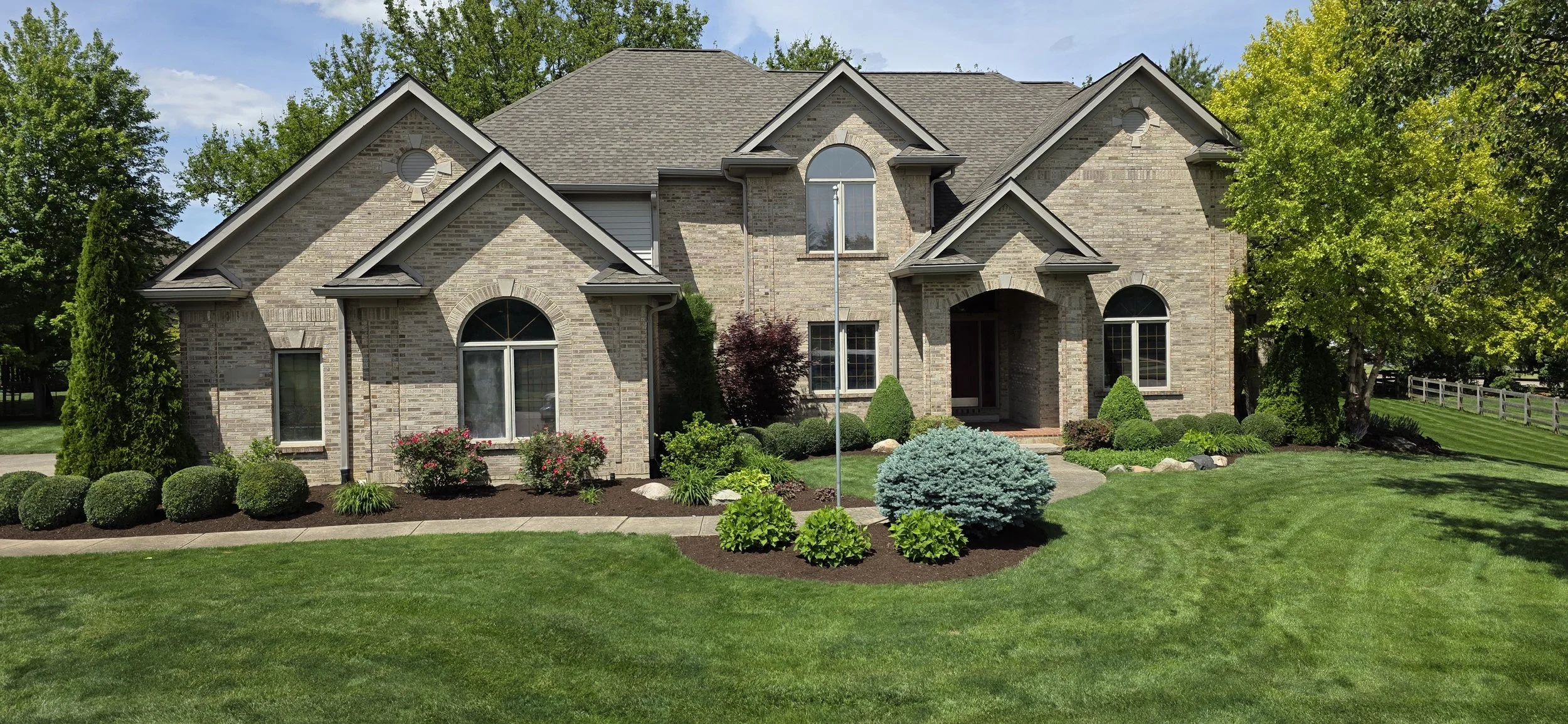 A large brick house with multiple gabled roofs, arched and rectangular windows, surrounded by a well-manicured lawn, bushes, trees, and flower beds under a partly cloudy sky.