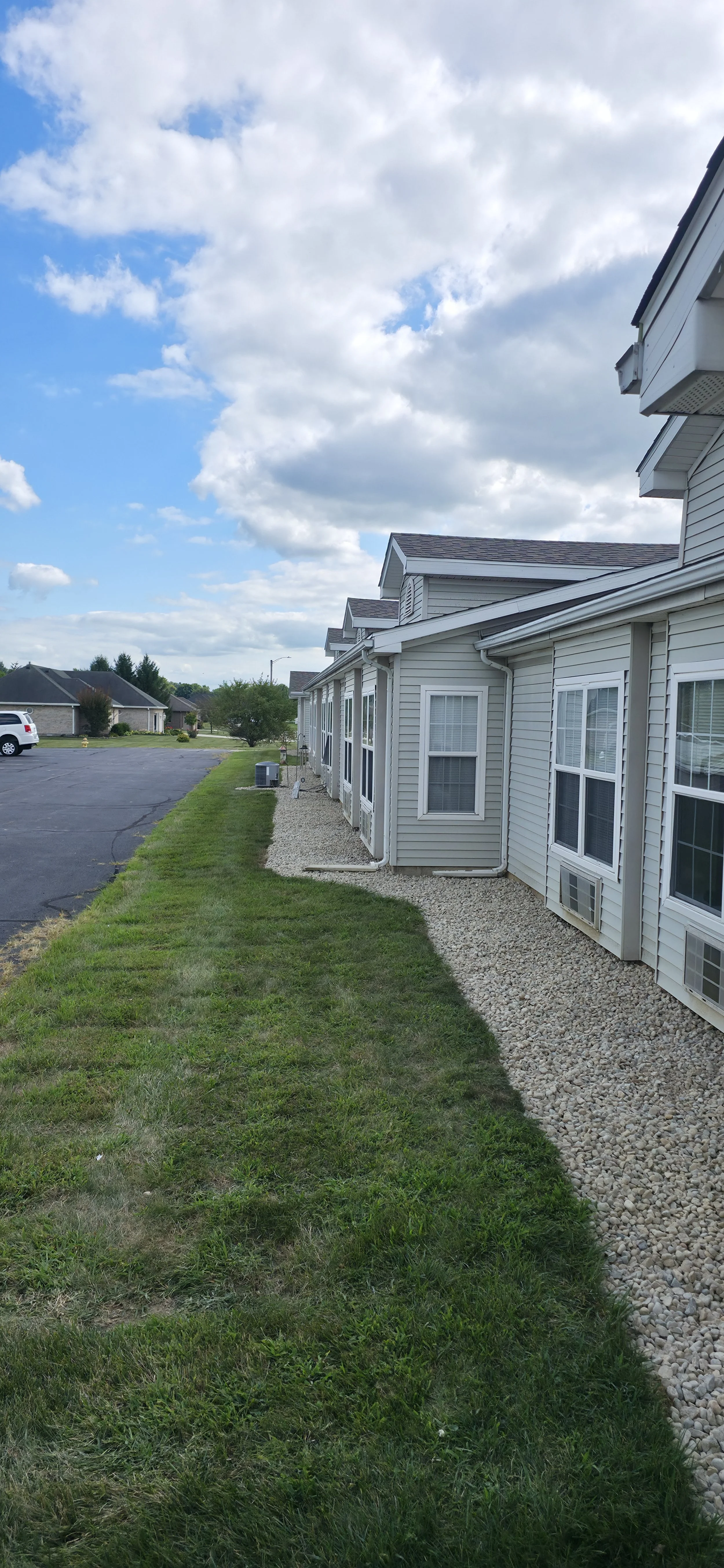 Apartment complex with grass lawn, gravel walkways, and parking lot under a partly cloudy sky.