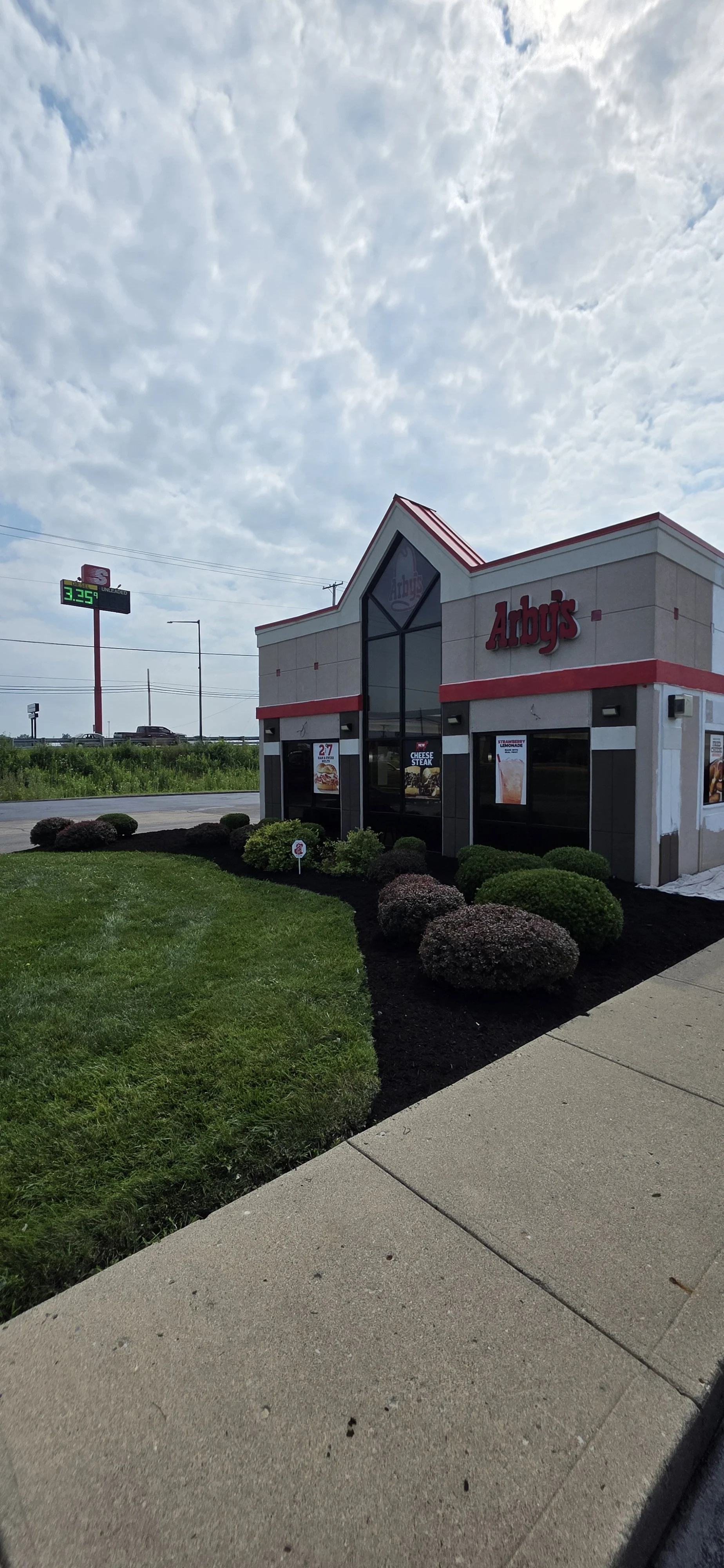 Exterior view of an Arby's fast food restaurant with landscaping, cloudy sky, and a gas station in the background.