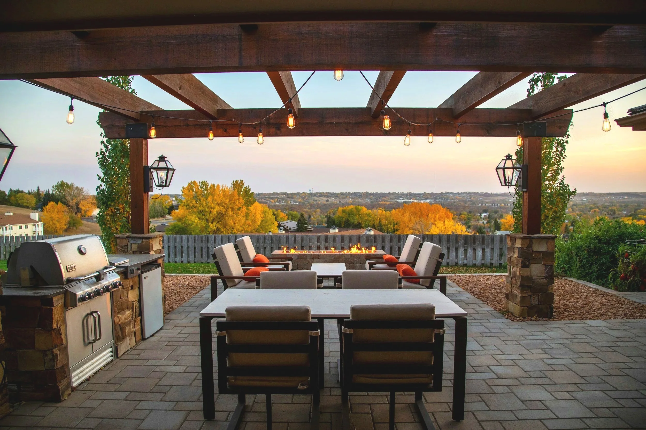 Outdoor patio with dining area, fireplace, string lights, barbecue grill, view of trees with autumn foliage, wooden pergola, and suburban landscape in the background.
