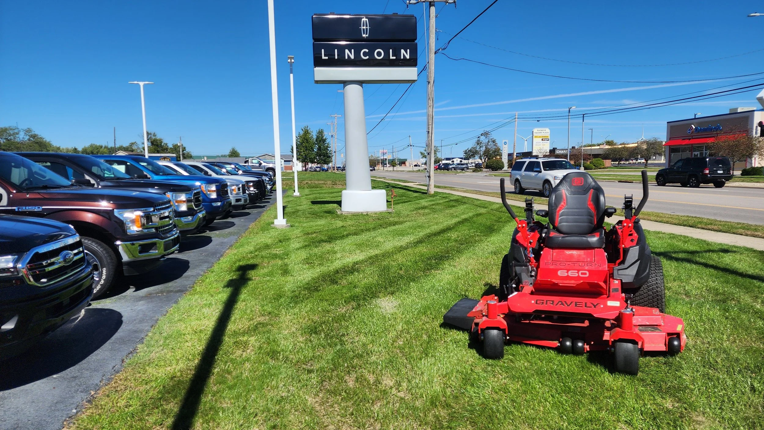 A red Gravely zero-turn lawn mower parked on a patch of grass near a Lincoln dealership sign, with a row of black and blue trucks lined up beside the dealership in bright sunshine.
