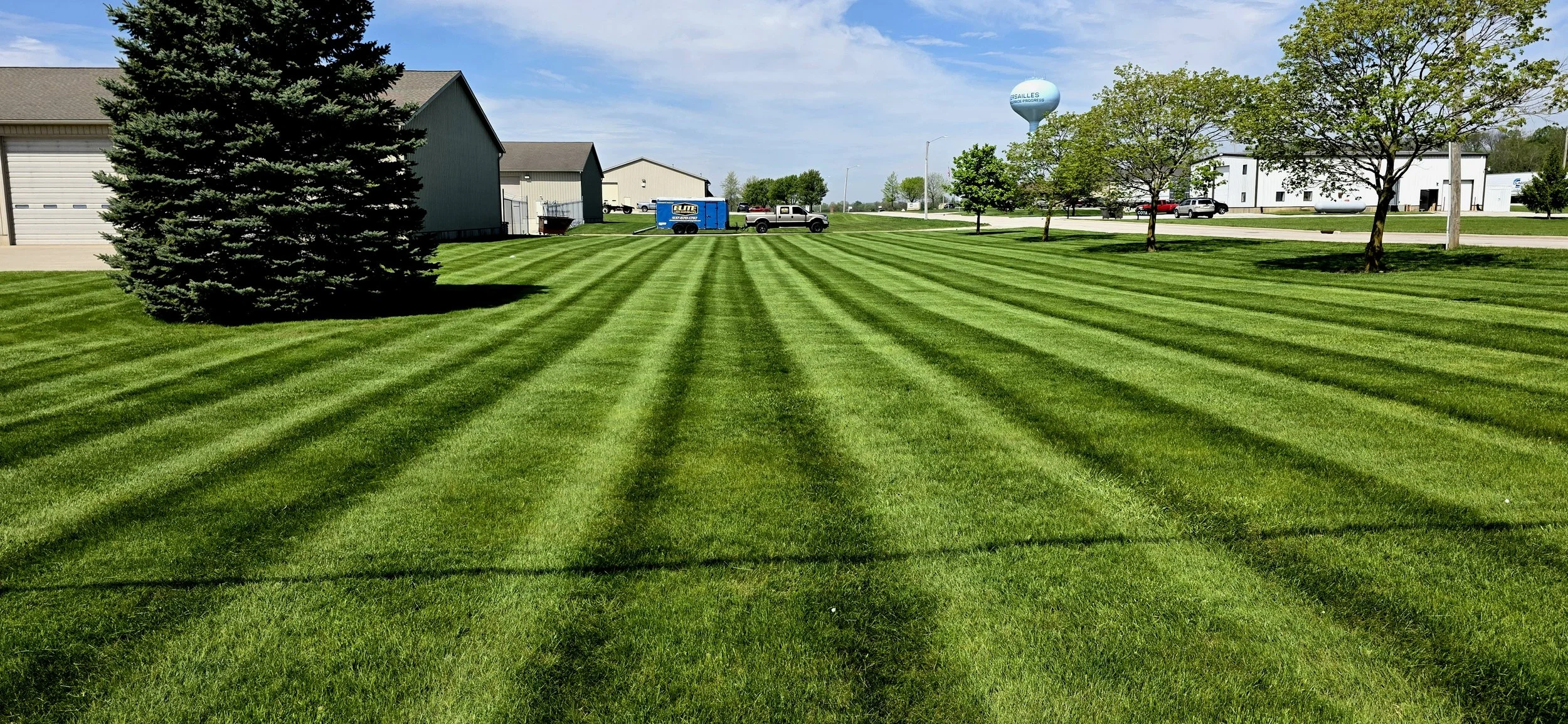 Well-maintained green lawn with striped grass and a few trees, bordered by buildings and parked vehicles under a partly cloudy sky.