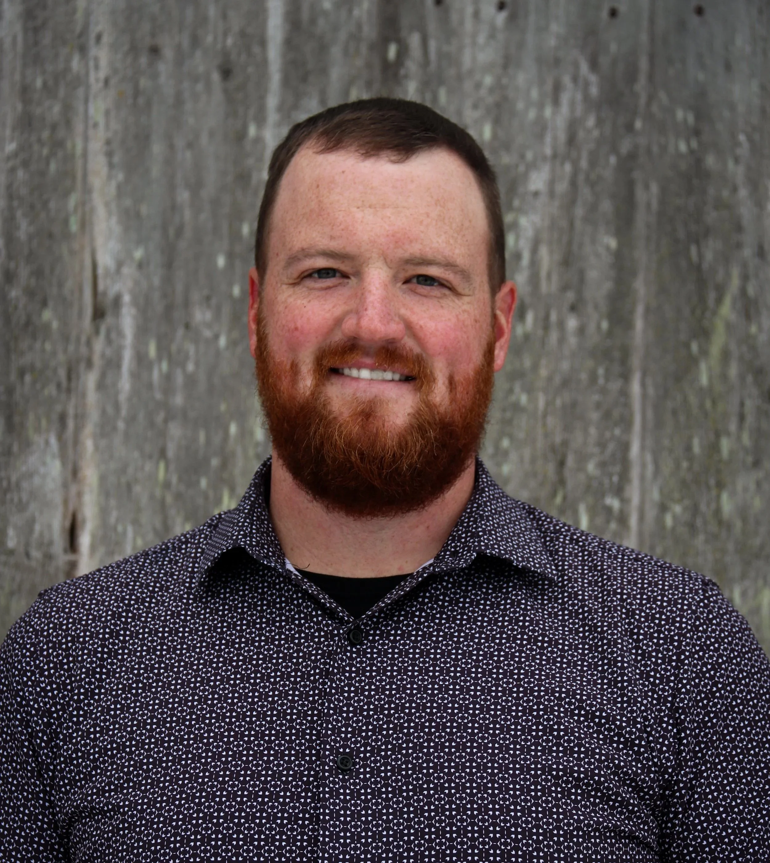 A man with a red beard and short brown hair, smiling, wearing a black collared shirt with a white pattern, standing outdoors in front of a wooden background.
