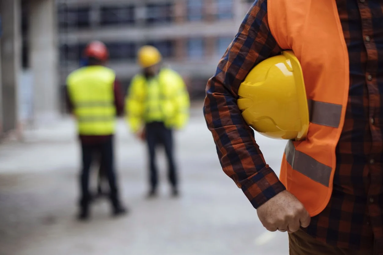 A construction worker holding a yellow safety helmet standing on a construction site with two other workers in the background.