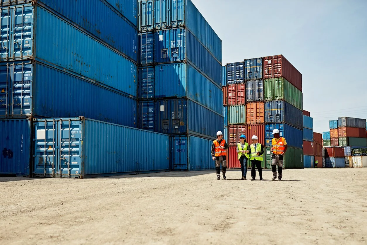 Four workers in safety vests and helmets walking and talking in a shipping yard with large stacked cargo containers.